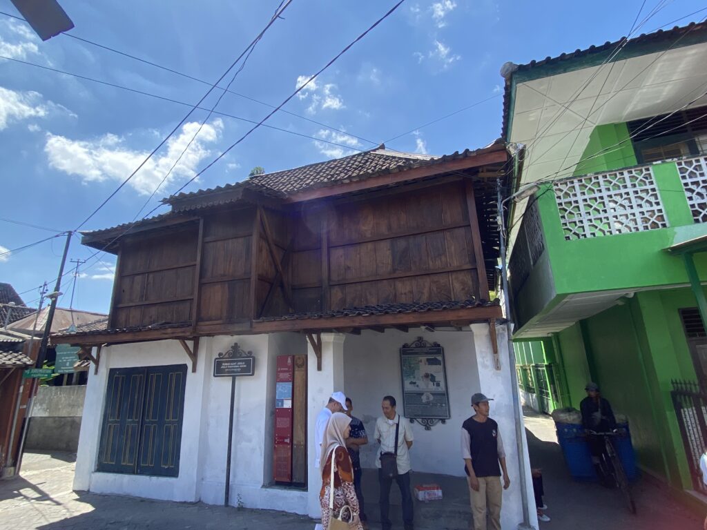 IMG_1657 - Mabur.co Street scene showing a white storefront with a dark wooden upper building, a bright green building on the right, and overhead power lines against a blue sky; several people stand near the entrance on a sunny day.