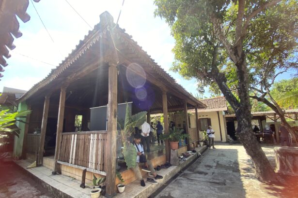 Open-air wooden pavilion with decorative eaves, people standing beneath, sun glare in a bright courtyard with potted plants nearby.