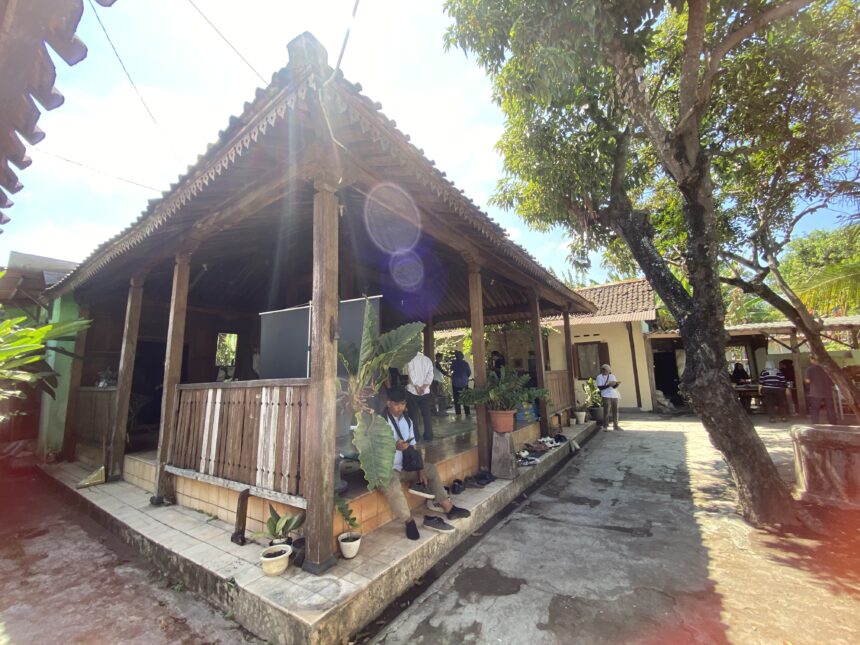Open-air wooden pavilion with decorative eaves, people standing beneath, sun glare in a bright courtyard with potted plants nearby.
