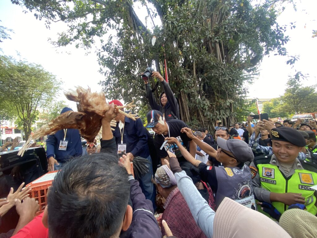 Crowd of people outdoors at a festival-like event, someone holds a large brown chicken above the crowd while photographers film.