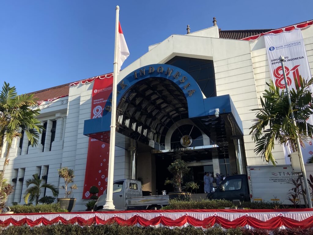 IMG_1765 - Mabur.co White modern bank building with a blue arched entrance, Indonesian flags, and red-white festive banners along the façade. palm trees frame the scene.