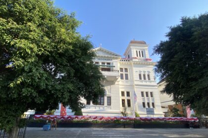White, ornate building partially visible behind green trees against a bright blue sky; the word 'INDONESIA' appears on the facade.