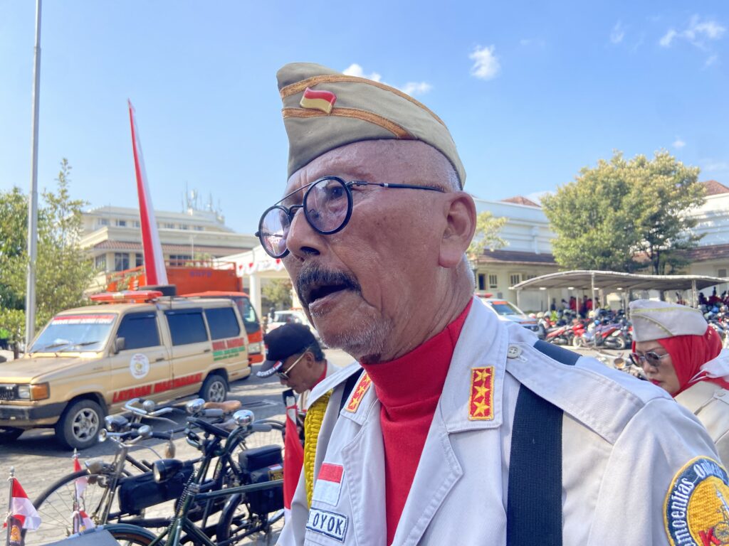 IMG_1793 - Mabur.co Close-up of a uniformed man with a beige ceremonial hat and glasses at an outdoor event, with a parade scene and emergency vehicles in the background.