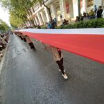 A long red-and-white banner stretched across a city street during a parade, with spectators on both sides watching from the sidewalk.