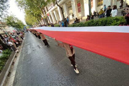 A long red-and-white banner stretched across a city street during a parade, with spectators on both sides watching from the sidewalk.