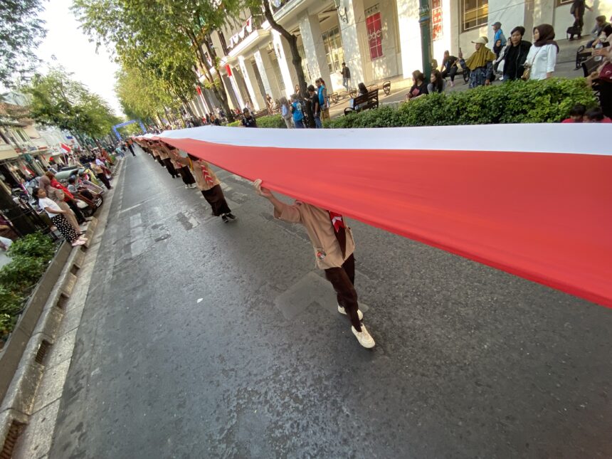 A long red-and-white banner stretched across a city street during a parade, with spectators on both sides watching from the sidewalk.