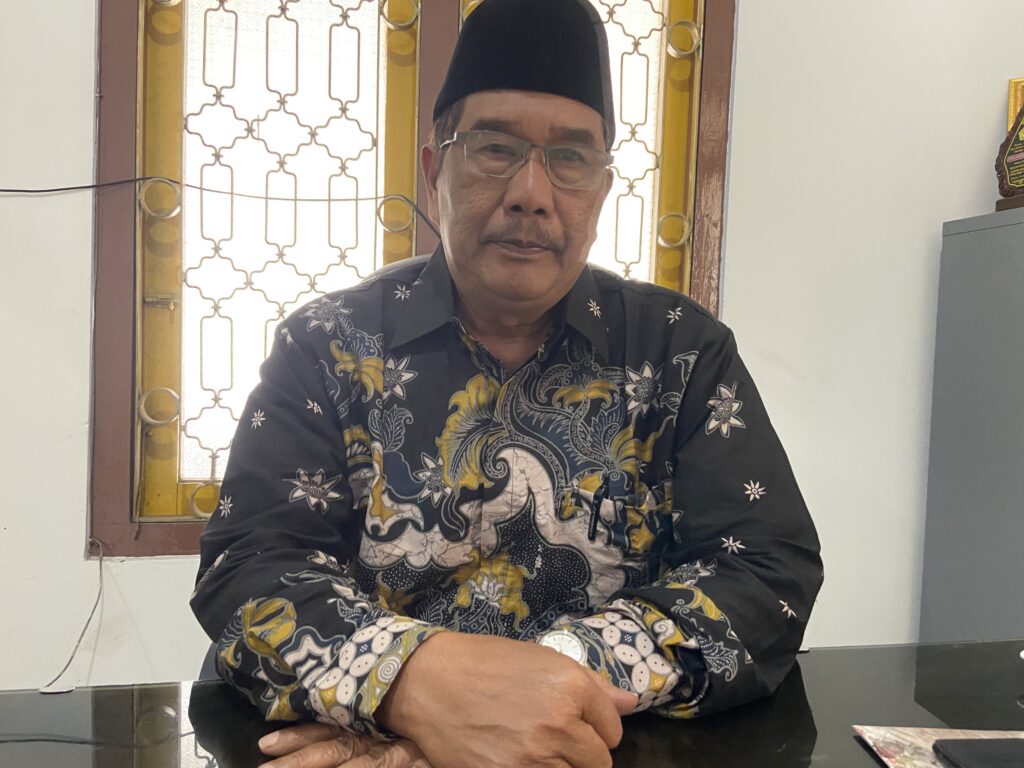 IMG_1881 - Mabur.co Man wearing a batik shirt and black peci hat sits at a glass table with folded hands, in front of a decorative window grille.