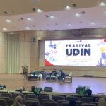 Speaker standing at a podium on a stage in a large conference hall, with seated panelists and a large banner that reads 'Festival Udin' behind them