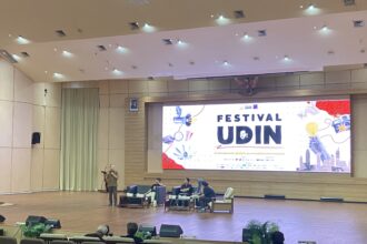 Speaker standing at a podium on a stage in a large conference hall, with seated panelists and a large banner that reads 'Festival Udin' behind them