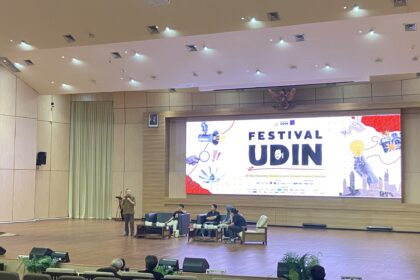 Speaker standing at a podium on a stage in a large conference hall, with seated panelists and a large banner that reads 'Festival Udin' behind them