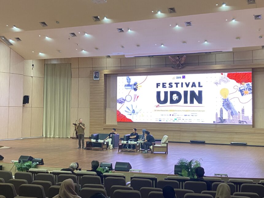 Speaker standing at a podium on a stage in a large conference hall, with seated panelists and a large banner that reads 'Festival Udin' behind them