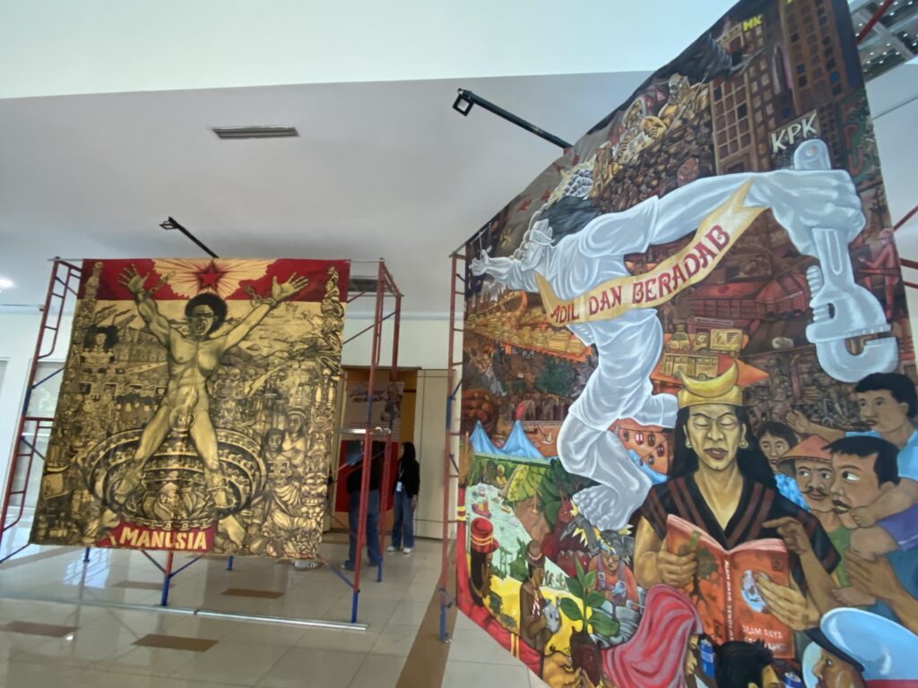 Indoor art installation with large colorful banners depicting historical scenes and people; visitors observe the displays in a gallery setting.
