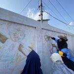 People in blue outfits painting a colorful mural on a white wall outdoors under a clear blue sky.