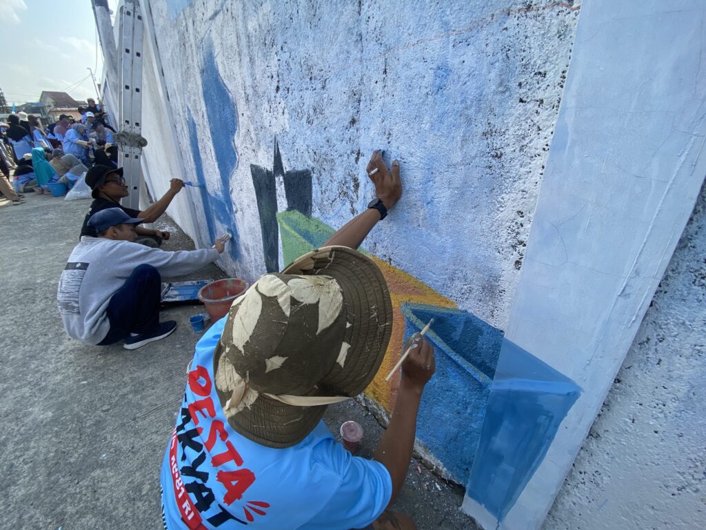 People painting a large blue mural along a wall, with several artists crouched or standing and using brushes and buckets of paint nearby.