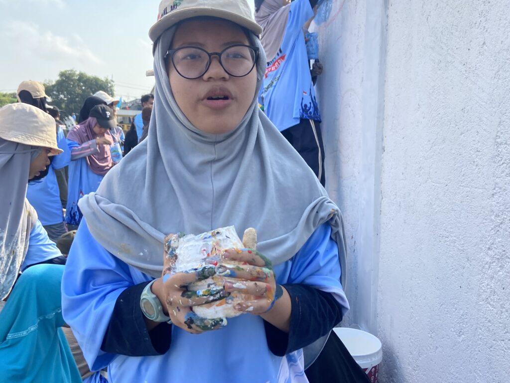 Woman wearing a hijab and glasses, holding a paint-splattered palette in an outdoor group painting activity with others in the background.