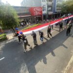 Parade on a city street with uniformed soldiers carrying a long red-and-white banner; onlookers on the sidewalk.