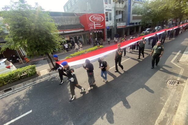 Parade on a city street with uniformed soldiers carrying a long red-and-white banner; onlookers on the sidewalk.