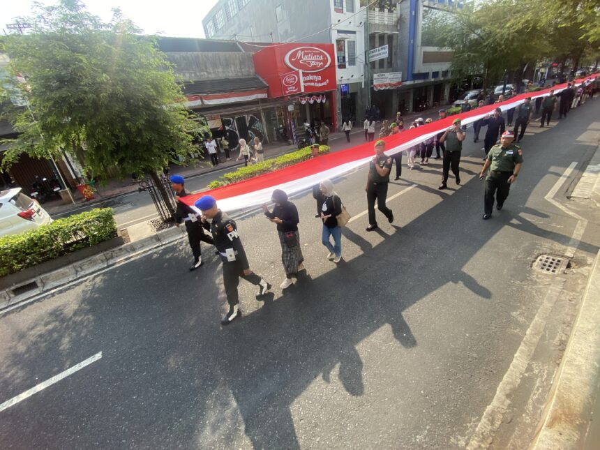 Parade on a city street with uniformed soldiers carrying a long red-and-white banner; onlookers on the sidewalk.