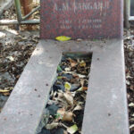 Red granite gravestone in a cemetery, with a sunken rectangular burial trough filled with fallen leaves, and the name 'A. Mangahaji' visible on the headstone.