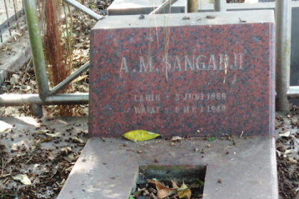 Red granite gravestone in a cemetery, with a sunken rectangular burial trough filled with fallen leaves, and the name 'A. Mangahaji' visible on the headstone.