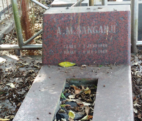 Red granite gravestone in a cemetery, with a sunken rectangular burial trough filled with fallen leaves, and the name 'A. Mangahaji' visible on the headstone.