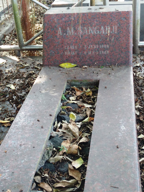 Red granite gravestone in a cemetery, with a sunken rectangular burial trough filled with fallen leaves, and the name 'A. Mangahaji' visible on the headstone.