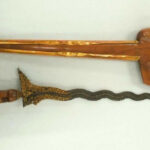 Ornate dagger with a wavy black blade and gold filigree guard beside a curved brown sheath.