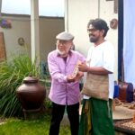 Two men stand and exchange a framed item as smiles abound; an elderly man in a flat cap hands it to a taller man in white shirt and green pants, in a garden setting.