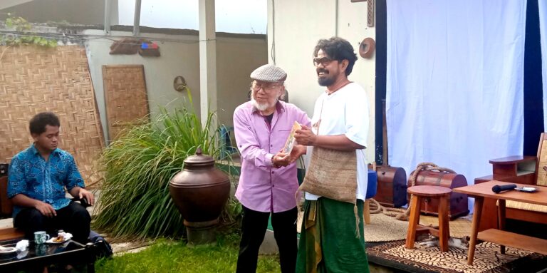 Two men stand and exchange a framed item as smiles abound; an elderly man in a flat cap hands it to a taller man in white shirt and green pants, in a garden setting.