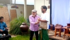 Two men stand and exchange a framed item as smiles abound; an elderly man in a flat cap hands it to a taller man in white shirt and green pants, in a garden setting.