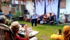 Woman in hijab speaks to an outdoor gathering holding papers, with people seated and taking photos in a garden setting.