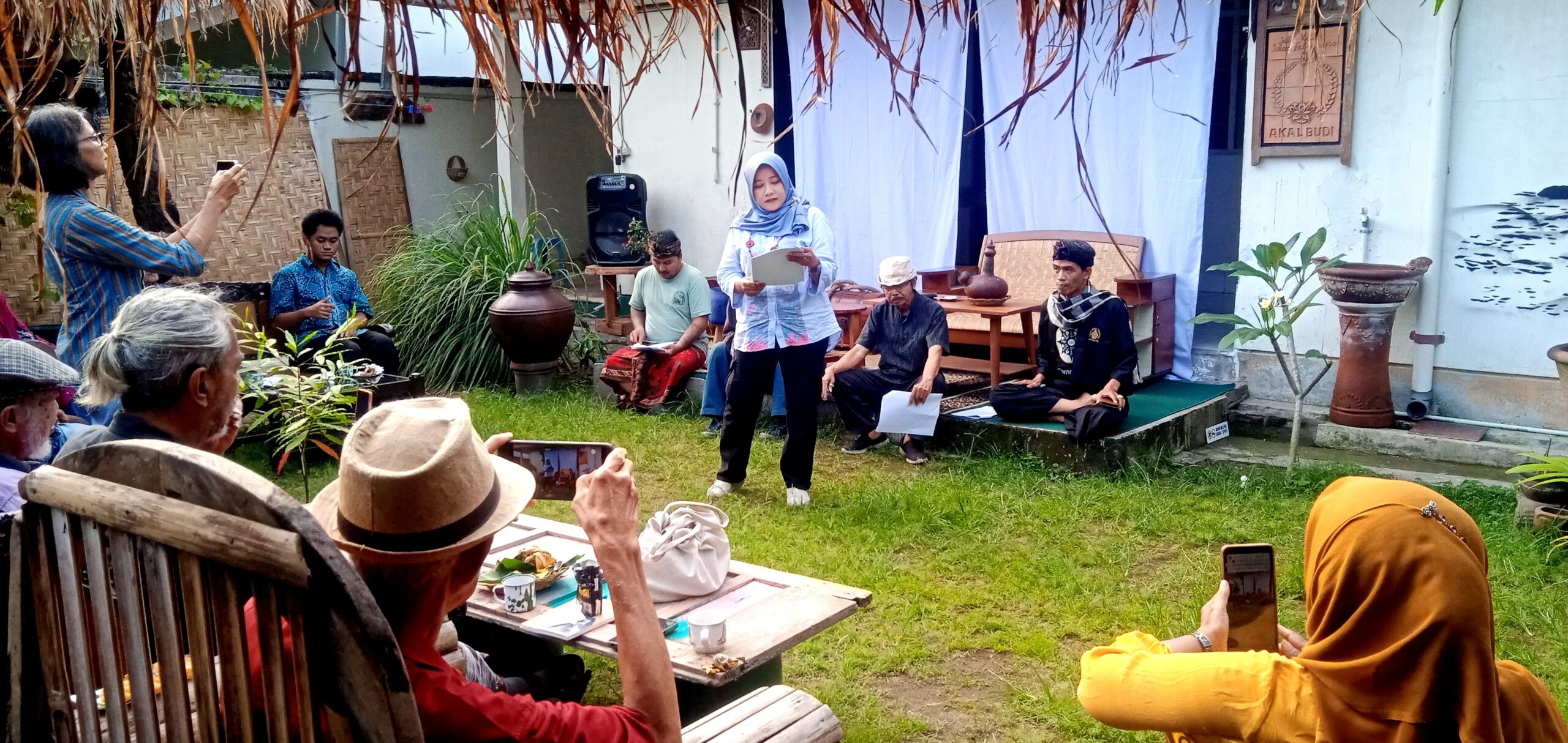 Woman in hijab speaks to an outdoor gathering holding papers, with people seated and taking photos in a garden setting.