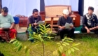 Four men sit on a wooden platform outdoors, reading from papers; a drum and plants are visible behind them.