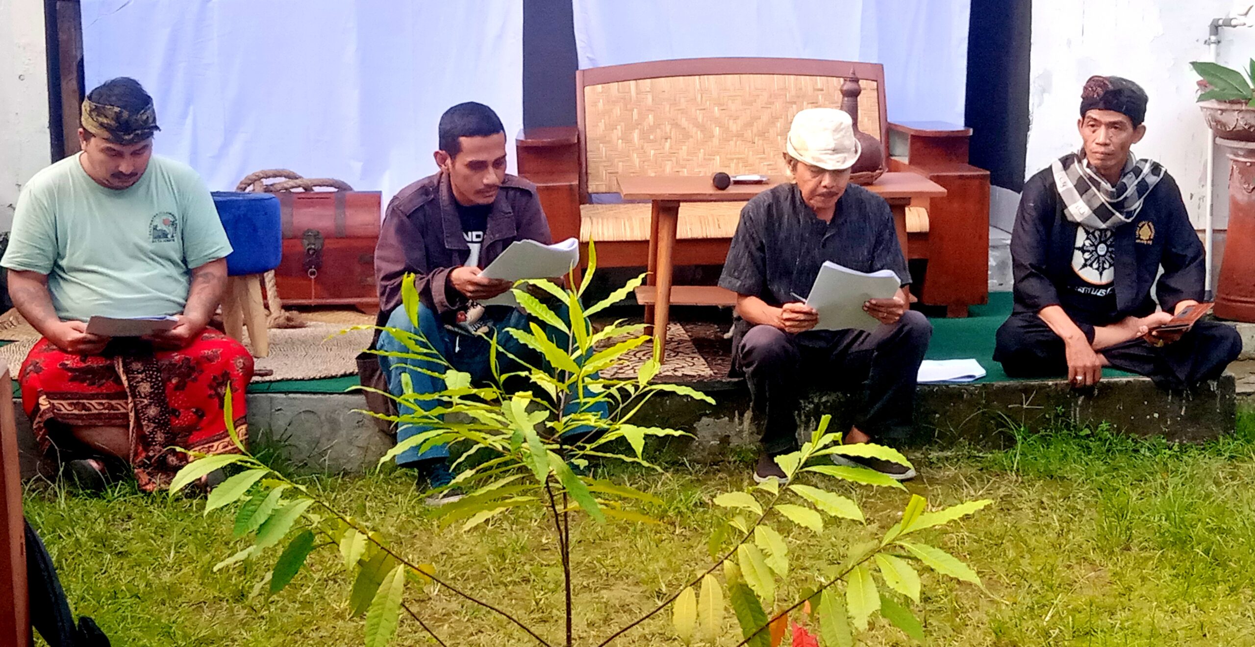 Four men sit on a wooden platform outdoors, reading from papers; a drum and plants are visible behind them.