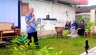 Older man in a blue shirt speaks into a handheld mic, reading from papers at an outdoor yard talk.