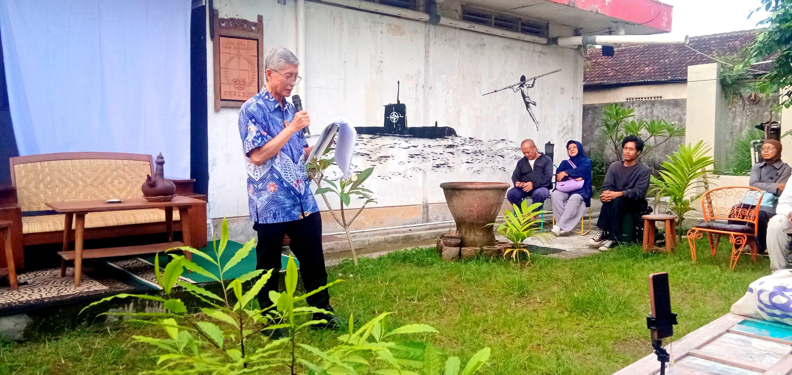 Older man in a blue shirt speaks into a handheld mic, reading from papers at an outdoor yard talk.
