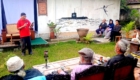 Man in a red shirt speaks into a microphone to a small outdoor audience in a garden setting.