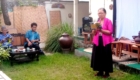Woman in a pink cardigan reads from a book into a microphone outdoors while two men listen nearby.