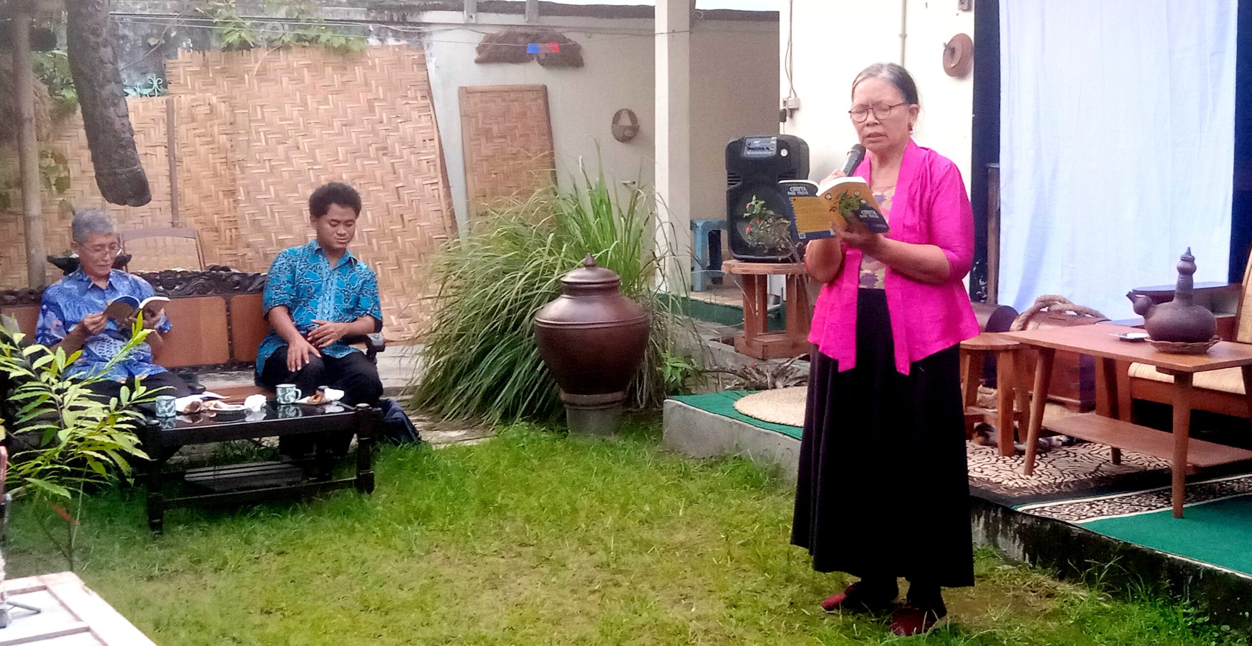 Woman in a pink cardigan reads from a book into a microphone outdoors while two men listen nearby.