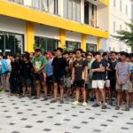 Large crowd of young men standing in a long queue outside a yellow-modern building while a man with a camera addresses them.