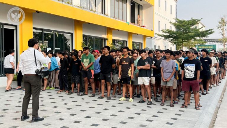 Large crowd of young men standing in a long queue outside a yellow-modern building while a man with a camera addresses them.