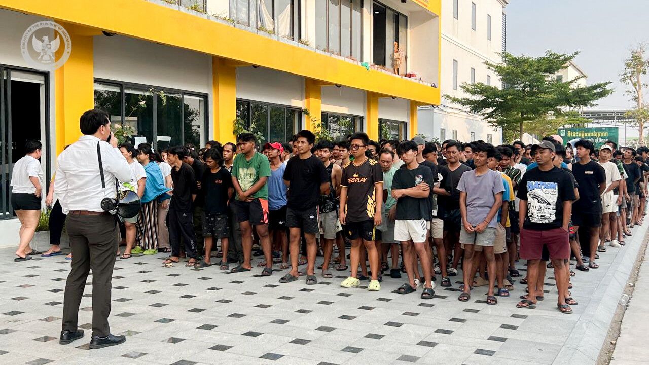 Large crowd of young men standing in a long queue outside a yellow-modern building while a man with a camera addresses them.