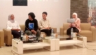 Panel discussion with four people seated on beige couches in a bright room; two women wearing hijabs and two men in casual clothes, rightmost woman holding papers.