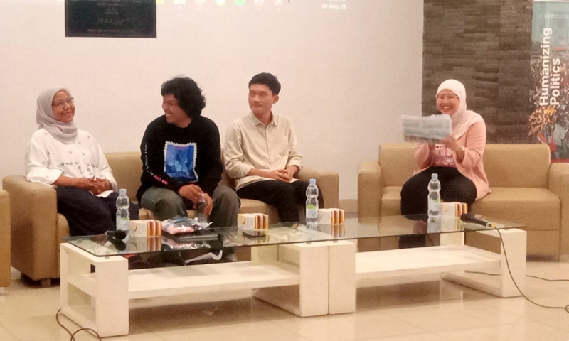 Panel discussion with four people seated on beige couches in a bright room; two women wearing hijabs and two men in casual clothes, rightmost woman holding papers.