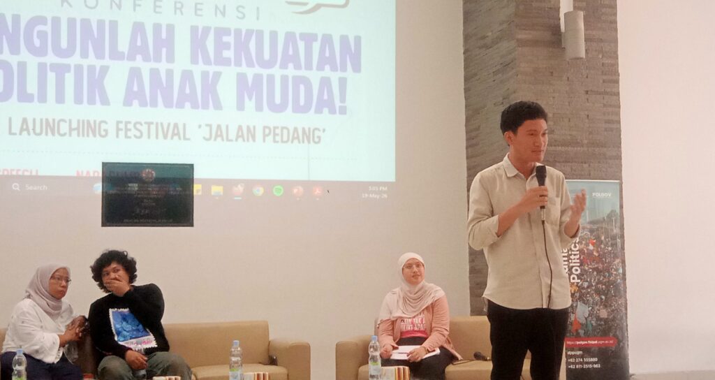 IMG_20260519_150523 - Mabur.co Man standing with a microphone presenting on a stage beside a large screen with event text in Indonesian, while seated panelists listen nearby.