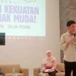 Man standing with a microphone presenting on a stage beside a large screen with event text in Indonesian, while seated panelists listen nearby.