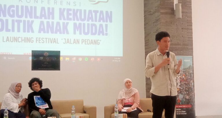 Man standing with a microphone presenting on a stage beside a large screen with event text in Indonesian, while seated panelists listen nearby.