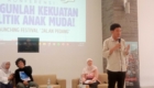 Man standing with a microphone presenting on a stage beside a large screen with event text in Indonesian, while seated panelists listen nearby.
