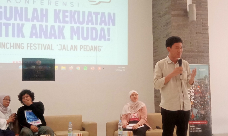 Man standing with a microphone presenting on a stage beside a large screen with event text in Indonesian, while seated panelists listen nearby.