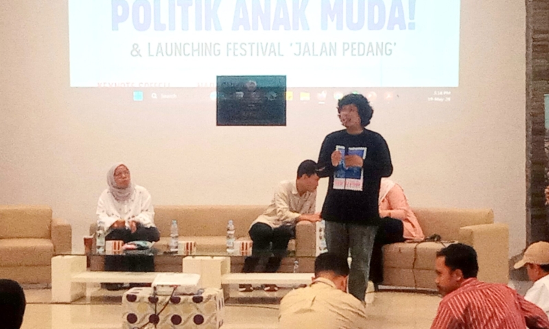 Panel discussion on a stage with three people seated and a speaker standing, projected slide reads POLITIK ANAK MUDA! & LAUNCHING FESTIVAL 'JALAN PEDANG'
